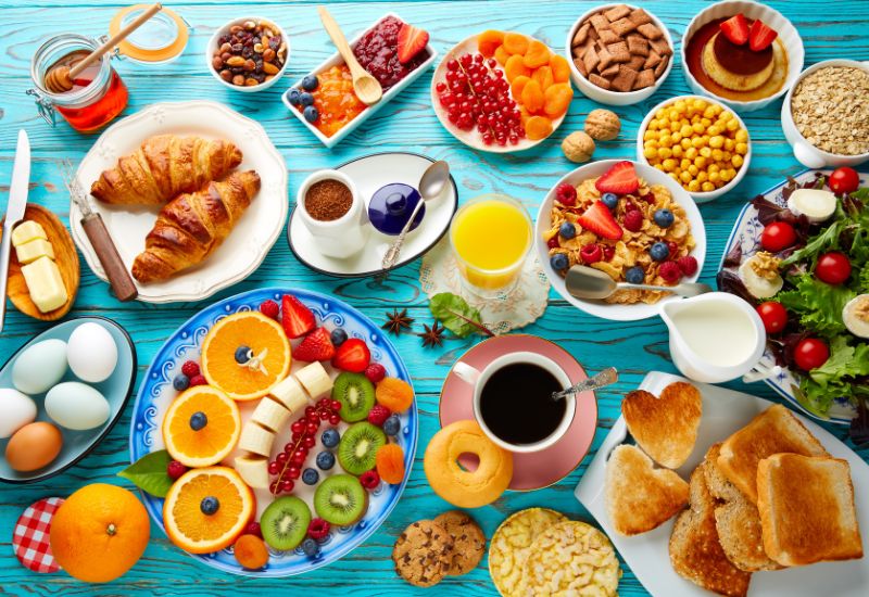 Různorodá zdravá snídaně na dřevěném stole, včetně ovocných smoothie, avokádového toastu, ovesné kaše, a chia pudingů