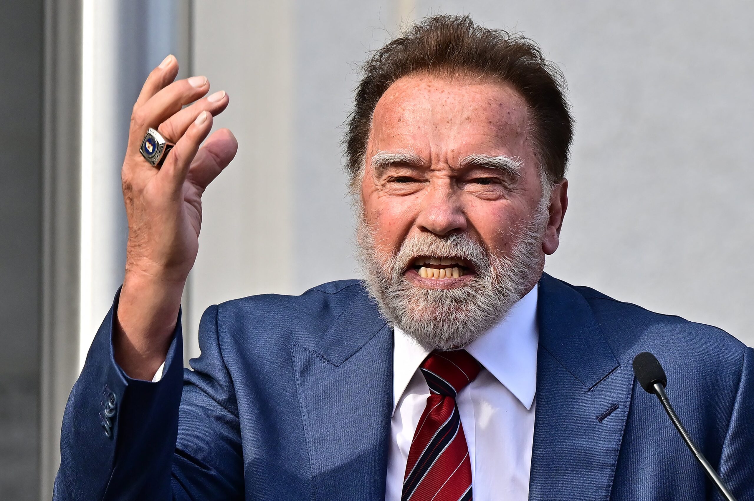 Arnold Schwarzenegger v obleku a kravatě, gestikulující při projevu na akci, viditelný mikrofon
