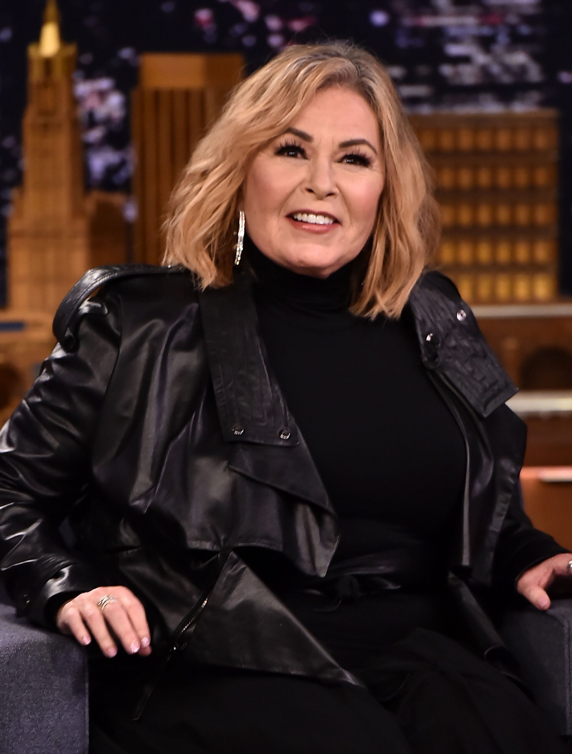 Osoba v černé kožené bundě a roláku sedí na talk show, usmívá se